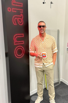 Paddy McGuinness standing next to a red ‘on air’ sign in a studio, wearing sunglasses, a patterned short-sleeve shirt and beige trousers, holding a red mug and smiling.