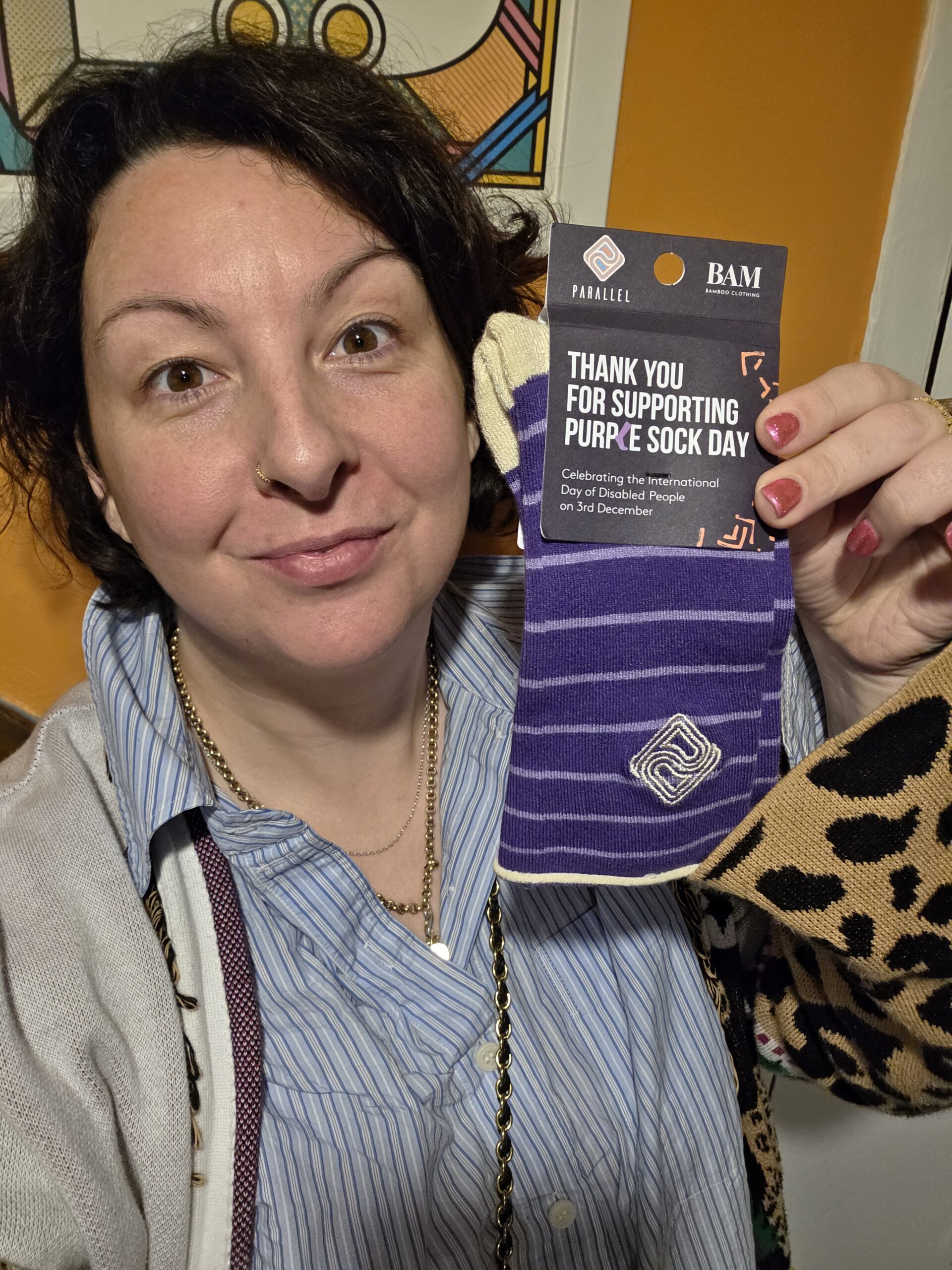 Person wearing Purpl socks draped around their shoulders while holding a ‘Sockfluencer’ card for Purple Sock Day 2025, promoting disability inclusion and awareness of long term health conditions.