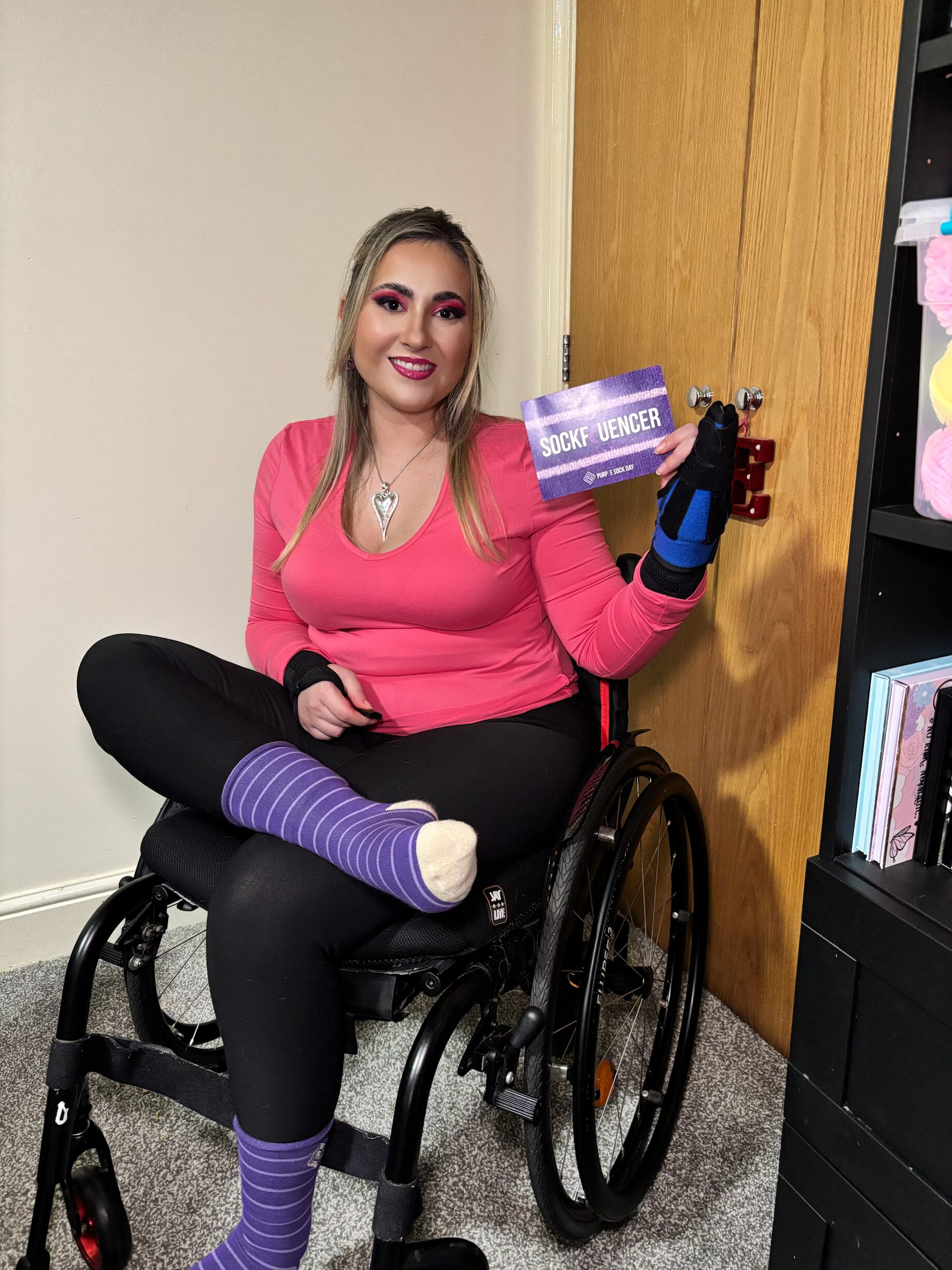 Person holding a pair of Purple Sock Day 2025 socks and smiling at the camera, supporting disability inclusion and chronic health condition awareness.