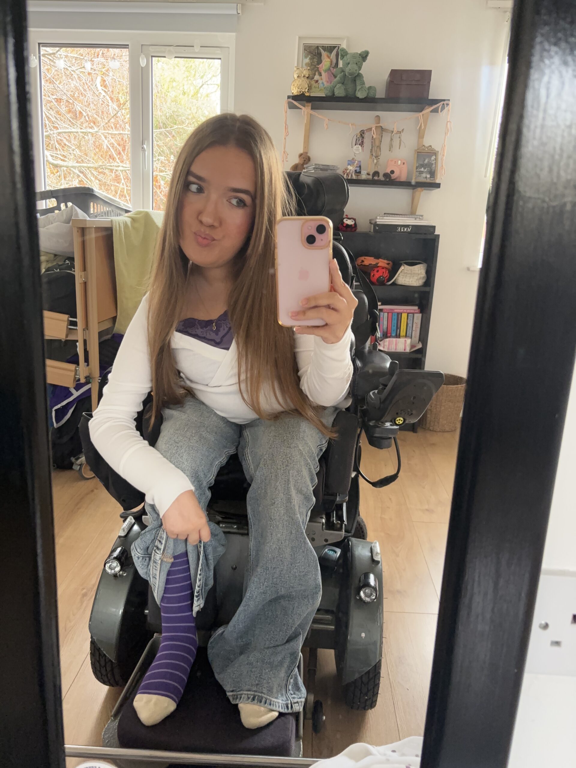 A young woman using a power wheelchair takes a mirror selfie while showing her purple striped Purpl socks for Purple Sock Day, supporting awareness of disabilities and long term health conditions and celebrating the inclusive Purpl discounts community.