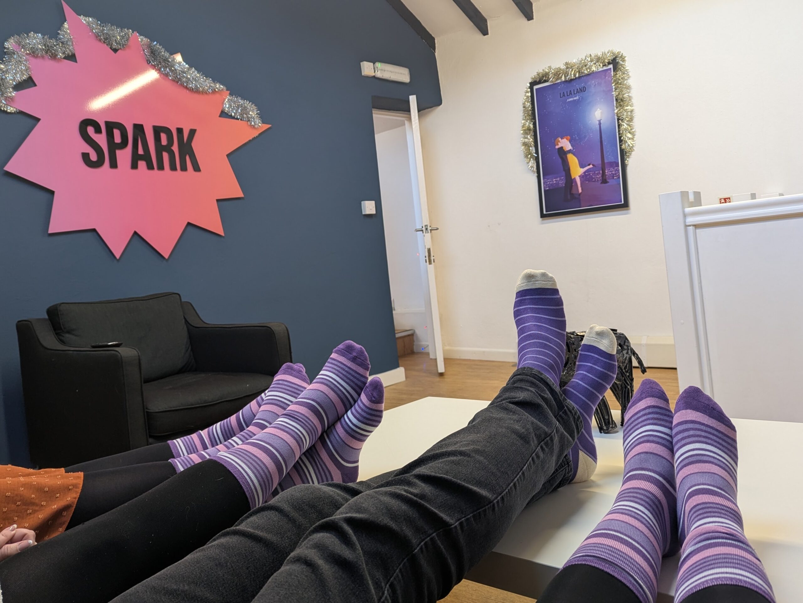 People wearing purple socks in an office with a SPARK sign and a La La Land poster, supporting Purple Sock Day to raise awareness for people living with disabilities and long term health conditions, alongside promoting inclusive savings through Purpl discounts, shopping discounts for disabled people, disabled travel discounts UK, and mobility discounts and offers.
