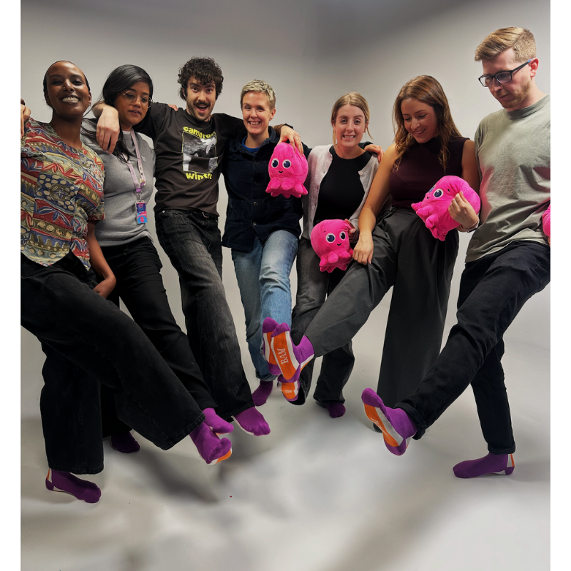 Group of people standing together holding pink octopus toys and wearing purple socks for Purple Sock Day, promoting awareness of disabilities and long-term health conditions and supporting accessibility and inclusion through Purpl discounts.