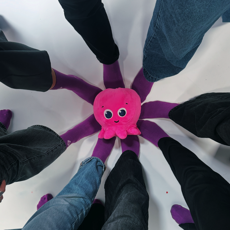 People standing in a circle with their purple socks pointed toward a pink plush octopus for Purple Sock Day, highlighting awareness of disabilities and long-term health conditions alongside accessibility and Purpl discounts.