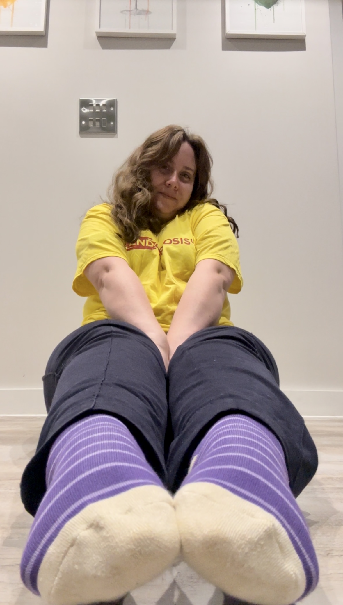 Person sitting on the floor wearing purple striped Purpl socks, smiling while supporting Purple Sock Day 2025; disability inclusion and chronic illness awareness.